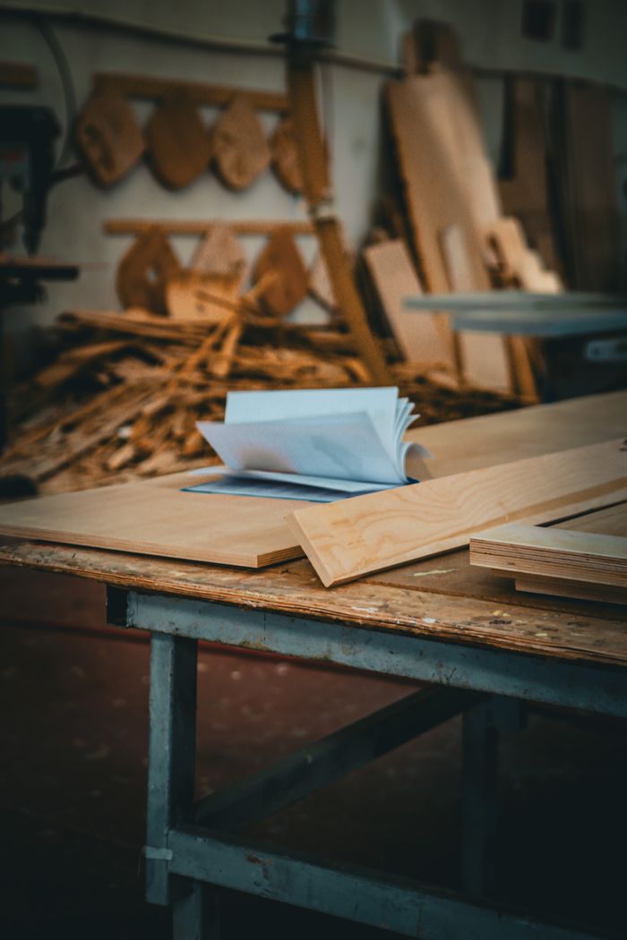 A detailed view of a carpentry table inside a workshop with stacks of wood and tools in the background.